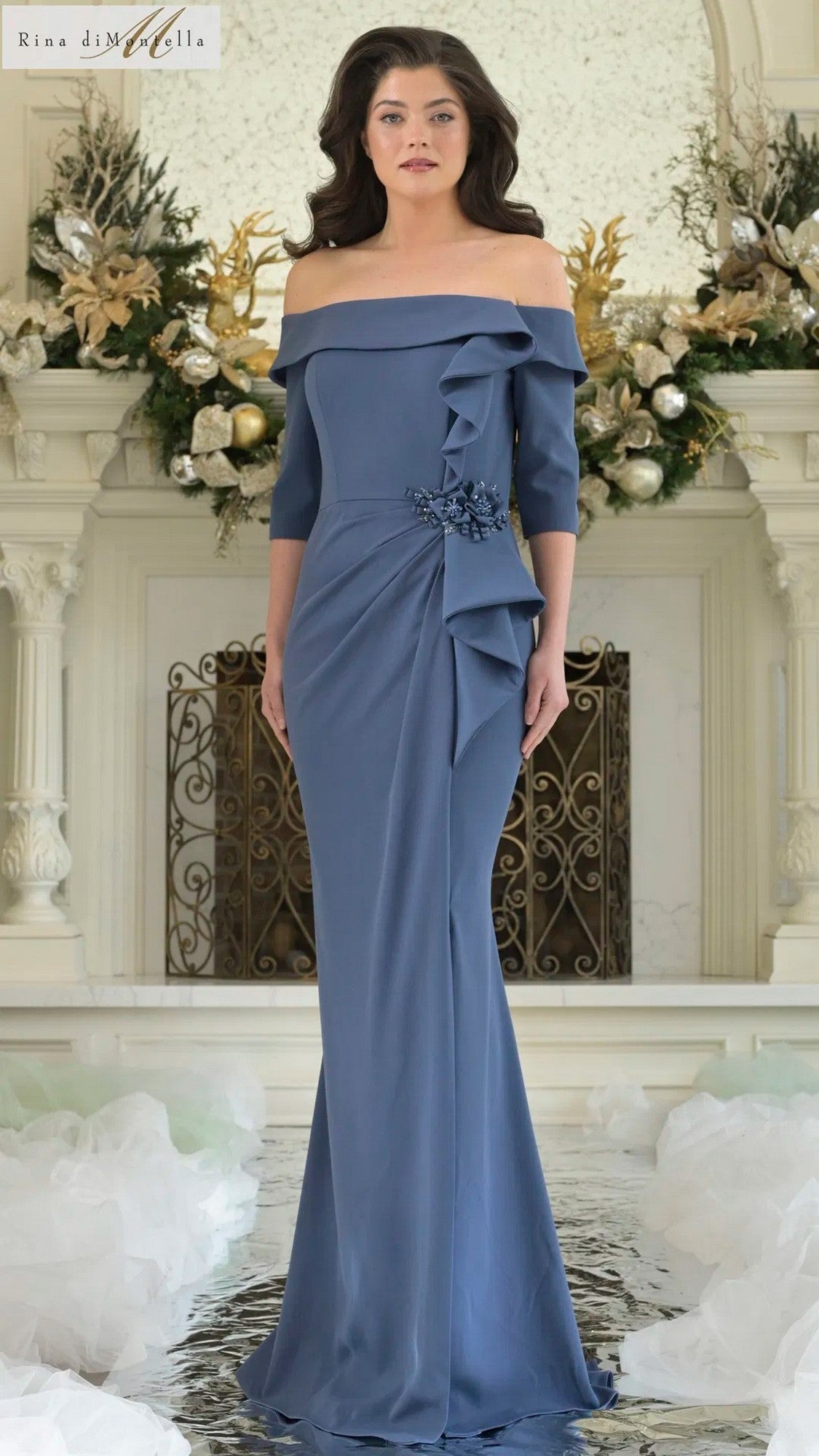 A woman stands indoors wearing a floor-length, deep royal Rina Di Montella RD3202 long formal dress, featuring off-the-shoulder style with floral embellishments and ruffle details, surrounded by ornate decor and floral arrangements.